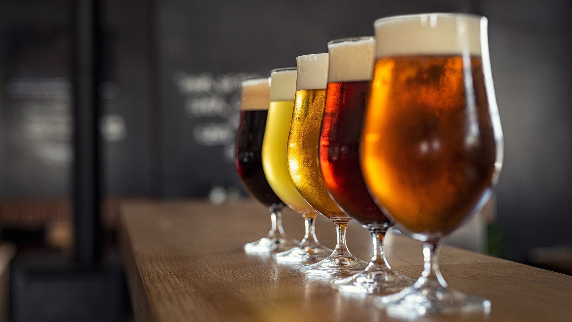 A row of assorted craft beers in stemmed glasses, ranging from pale gold to dark amber, lined up on a wooden bar.