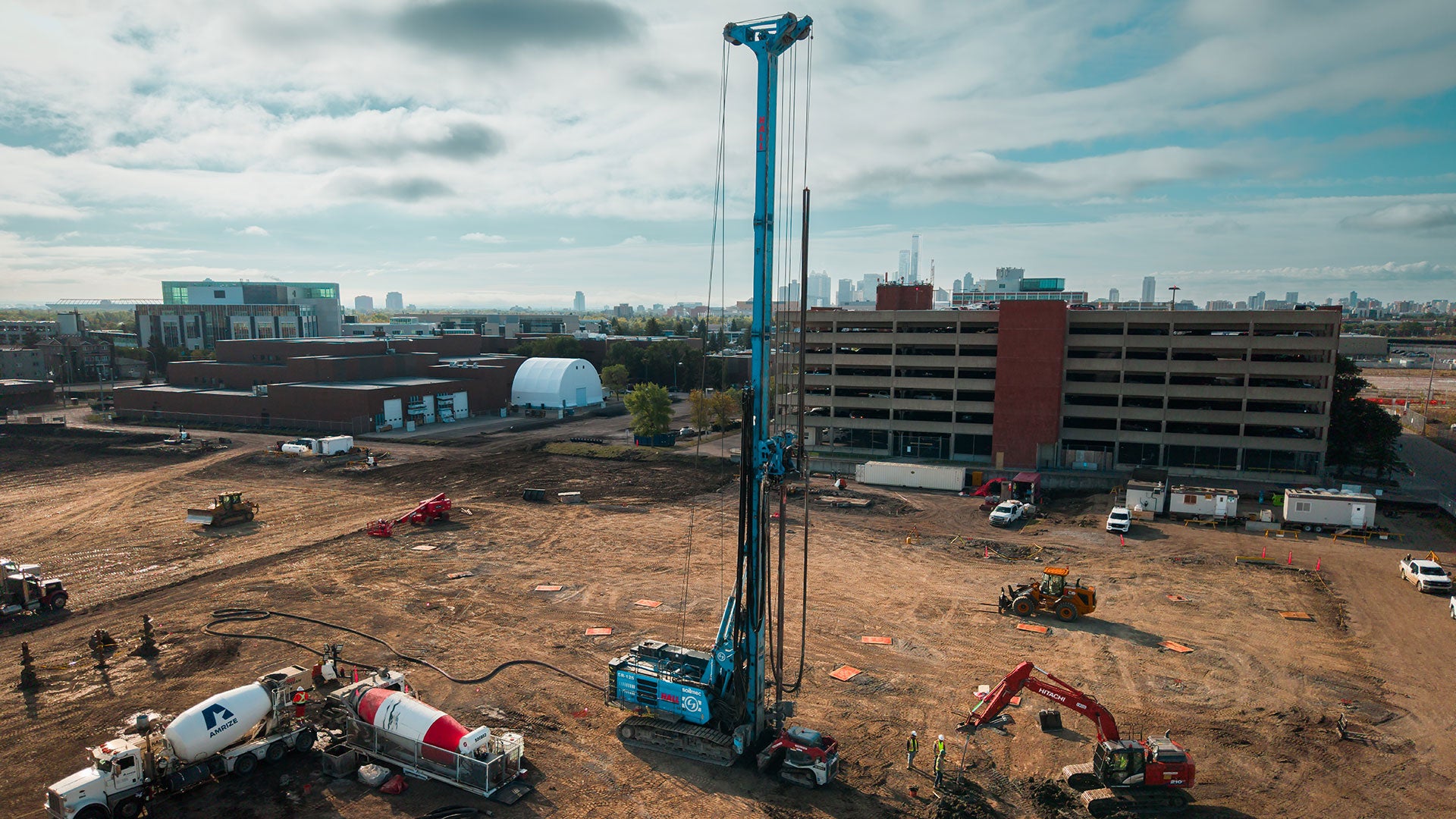 Construction site with a large drilling rig operating near a multi-level parking structure, surrounded by excavation equipment and building foundations.