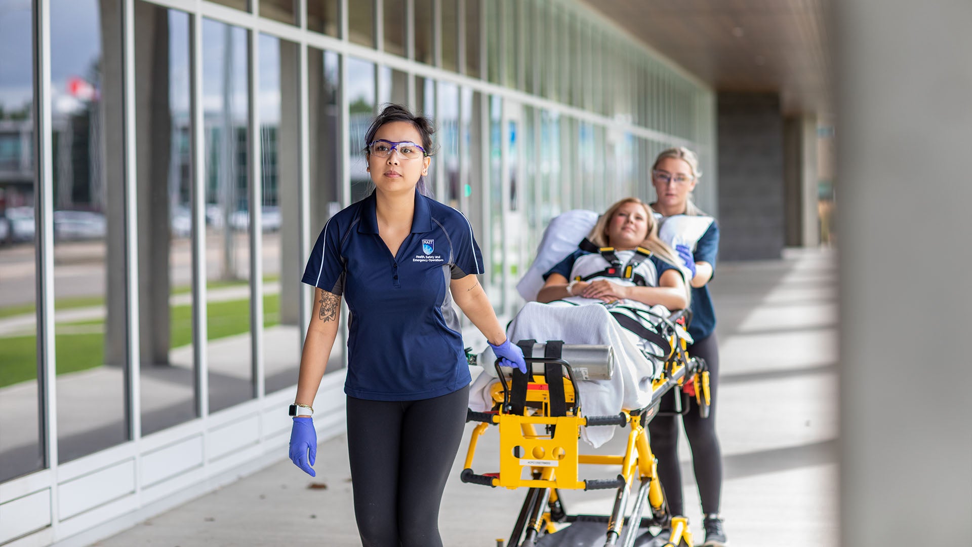 Two female Primary Care Paramedic students, one pulling a stretcher with a patient lying on it, while the other pushes. Both students are wearing rubber gloves and safety glasses.