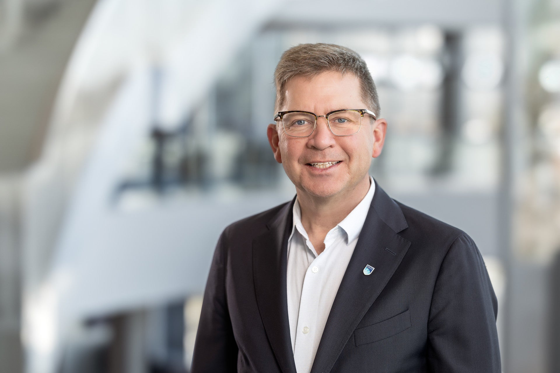Professional head-and-shoulders portrait of an individual wearing glasses, a white collared shirt and a dark business blazer with a lapel pin. The individual is positioned in the foreground of a modern indoor campus environment, with seating areas softly blurred in the background. Natural lighting and a shallow depth of field create a polished executive-style photograph, with the subject facing the camera in a professional setting.