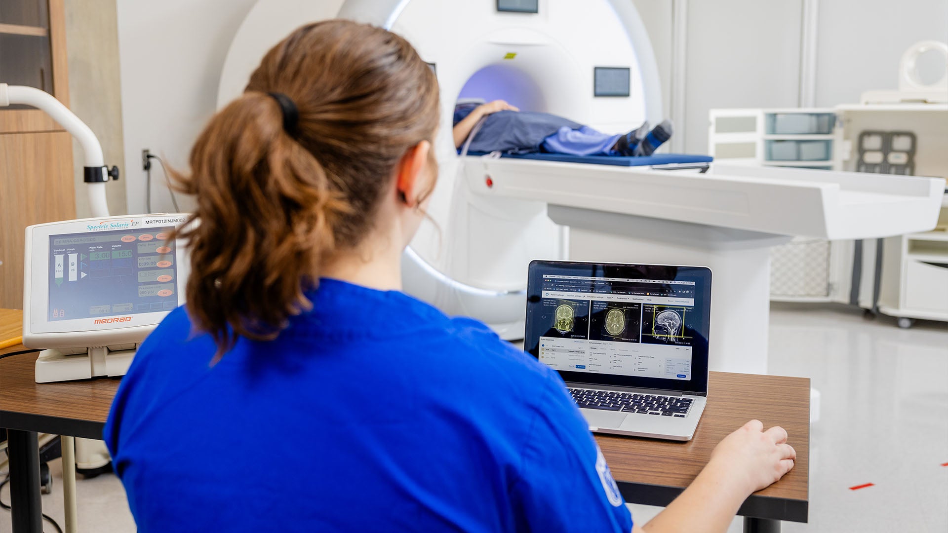 A student wearing blue scrubs looks at MRI results on a laptop while a patient lies inside an MRI machine.