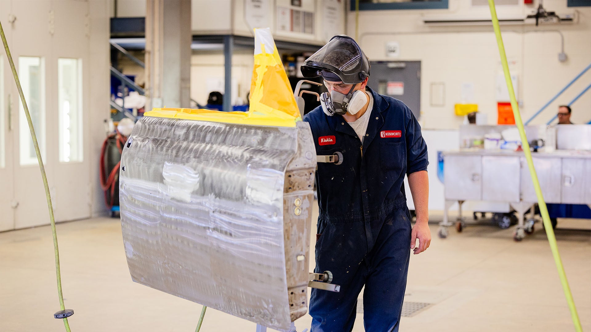 Person wearing a respirator and protective gear sanding or painting a vehicle panel in an automotive workshop.