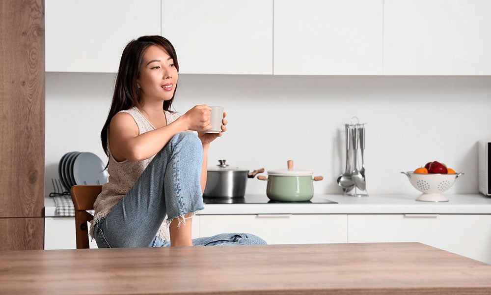 Person sitting on a kitchen counter holding a mug, with cookware and fruit visible in a bright modern kitchen.
