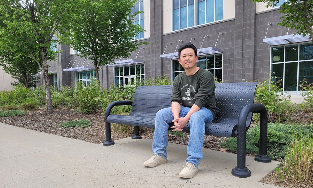 Person sitting on a black metal outdoor bench along a landscaped walkway near a modern building with large windows, wearing a green sweatshirt and jeans, with trees and shrubs in the background.
