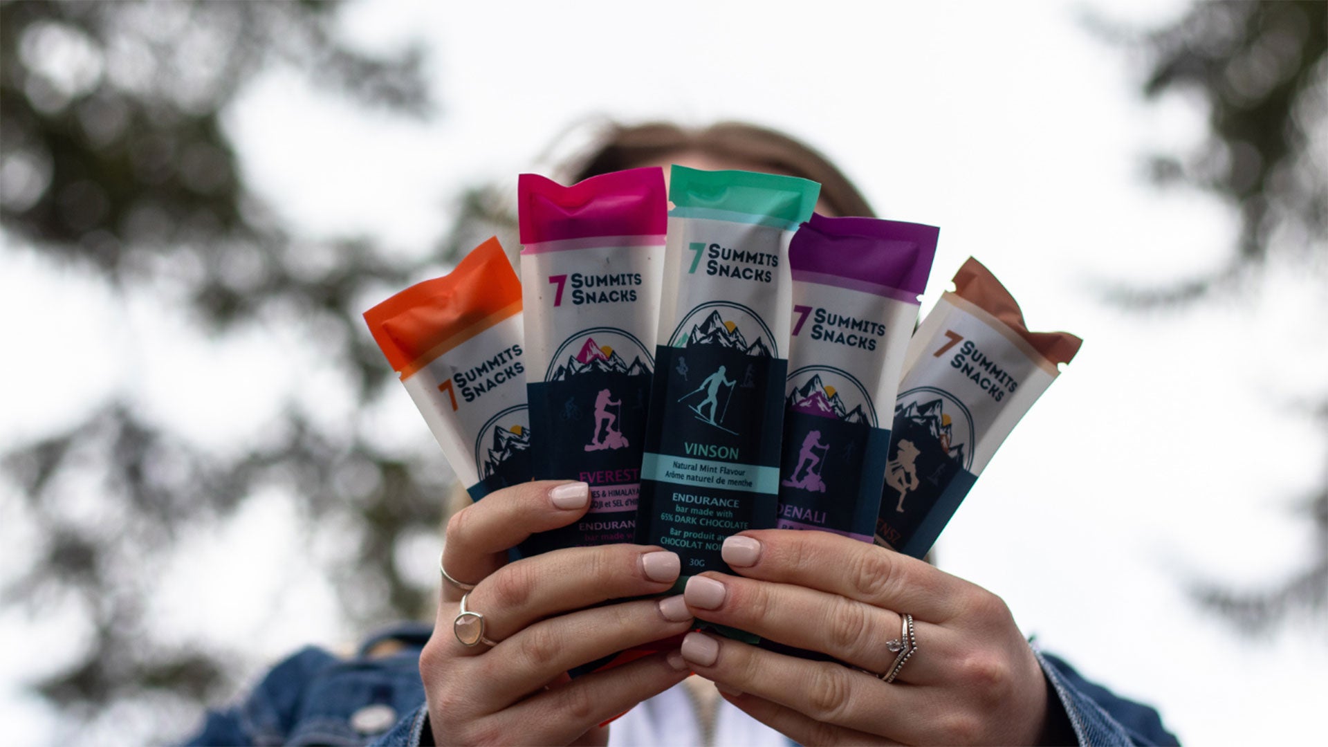 Person holding several colorful snack bar packages fanned out in both hands outdoors, with a blurred background of trees and visible product packaging in orange, green, purple, and white.