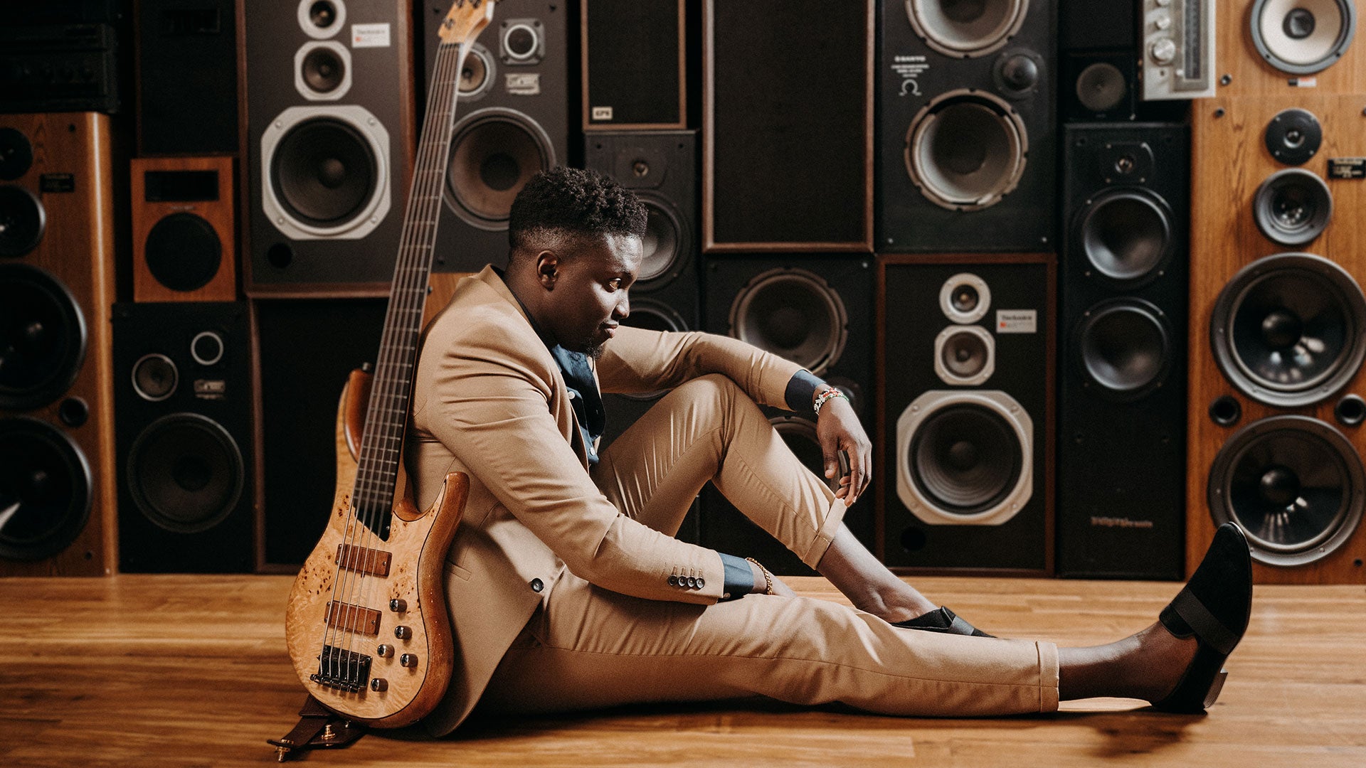 Person in a tan suit sitting on a wooden floor leaning against a wall of speakers while holding a bass guitar.
