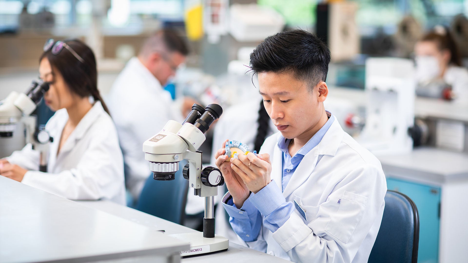 A group of dental technology students wearing white lab coats in a lab or classroom. Each student is sitting in front of a stereomicroscope. One student in the foreground is focused on examining a dental model.