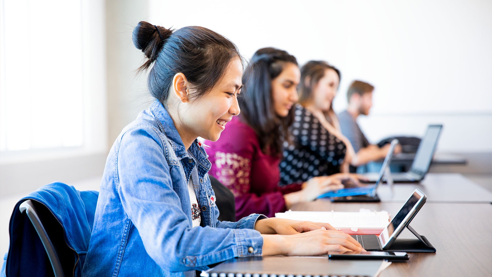 A female student attentively engaged in a classroom, using a tablet, with open books on the desk and peers working in the background. 