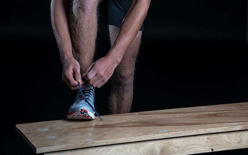 Person tying the laces of a running shoe while standing beside a wooden bench against a dark background.