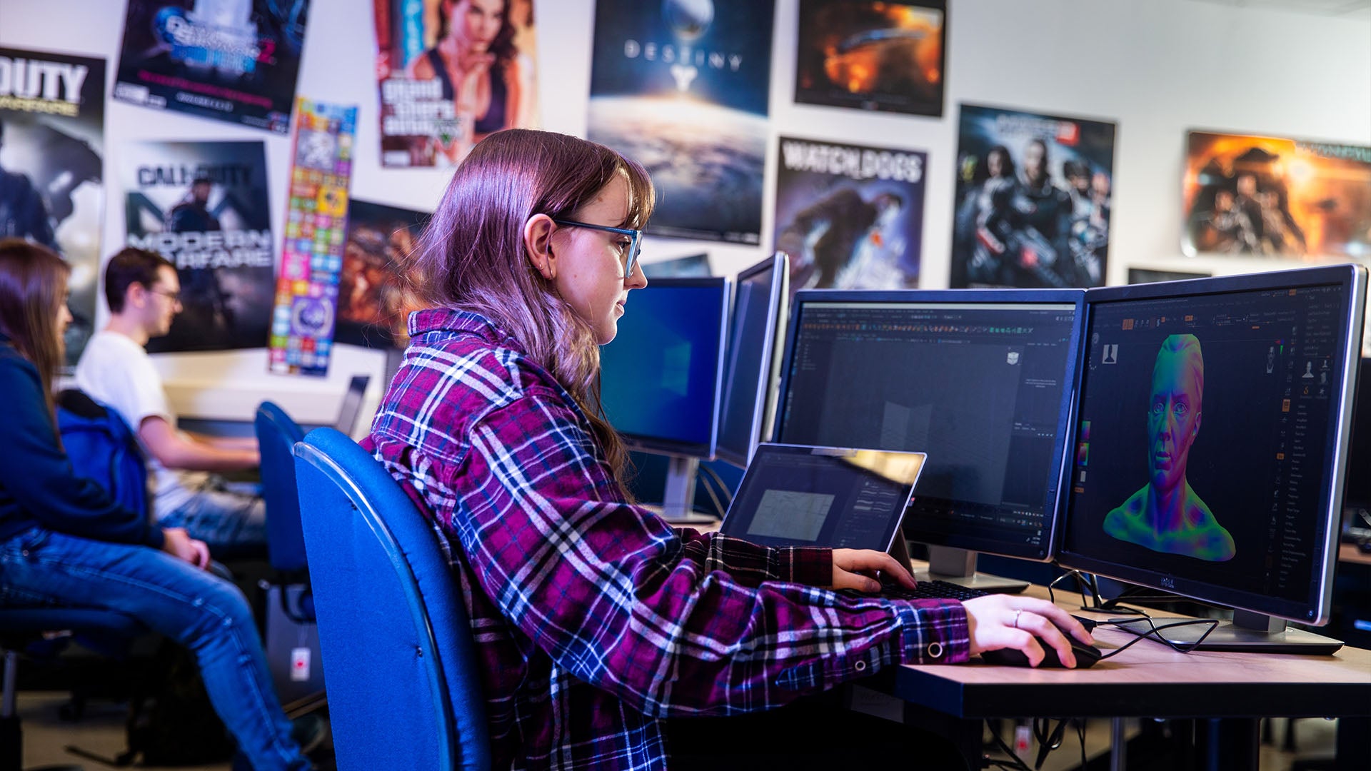 Woman working on 3D character model on a computer, surrounded by monitors and game posters in a classroom setting.