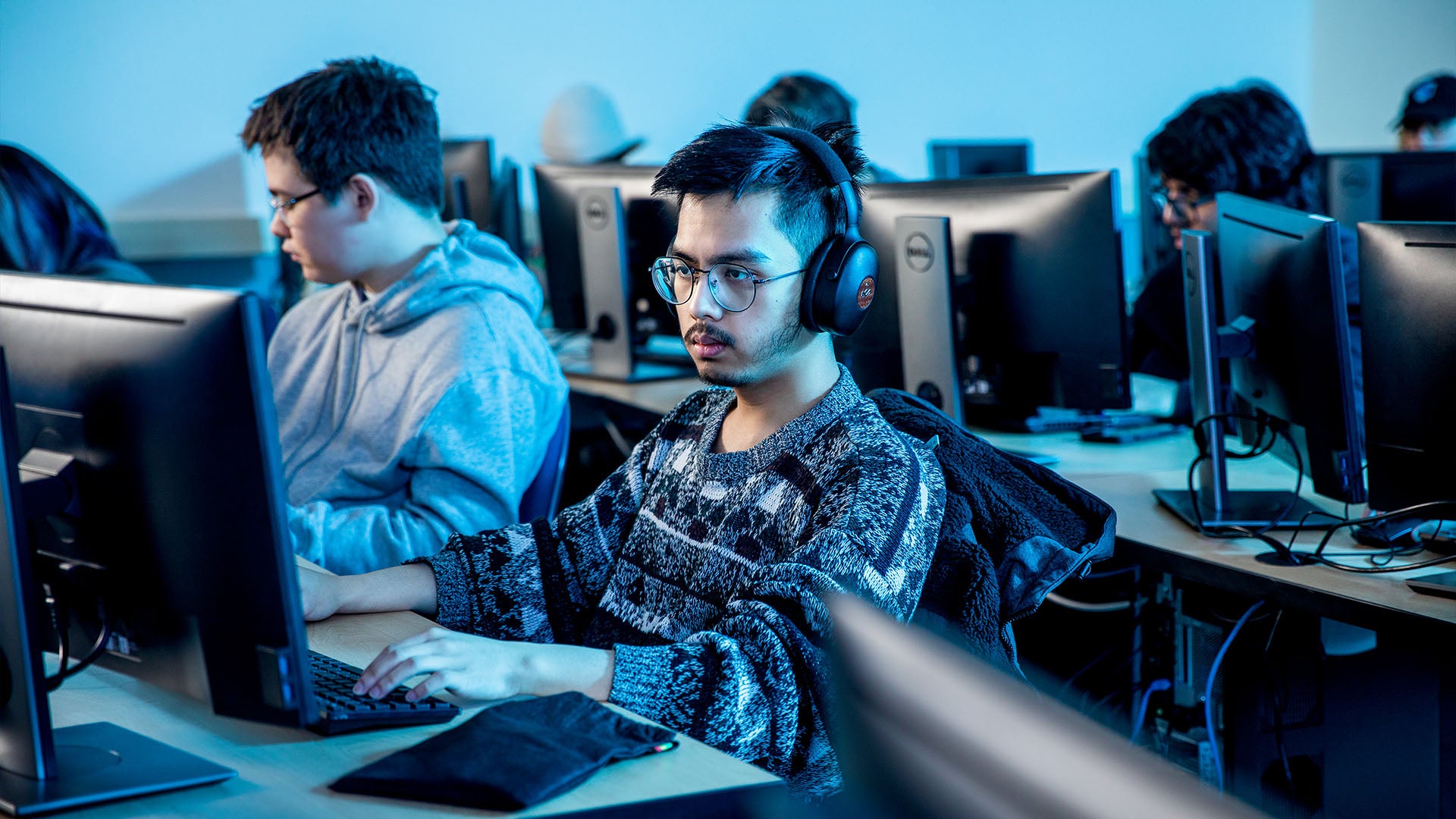 A male student wearing glasses and wireless headphones works on a desktop computer in a classroom, surrounded by other students engaged in their own work.