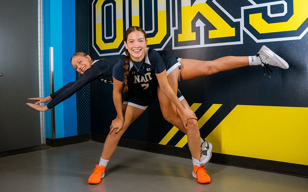 Two volleyball players in athletic uniforms posing playfully in front of a blue and gold Ooks-branded wall graphic inside a sports facility hallway.
