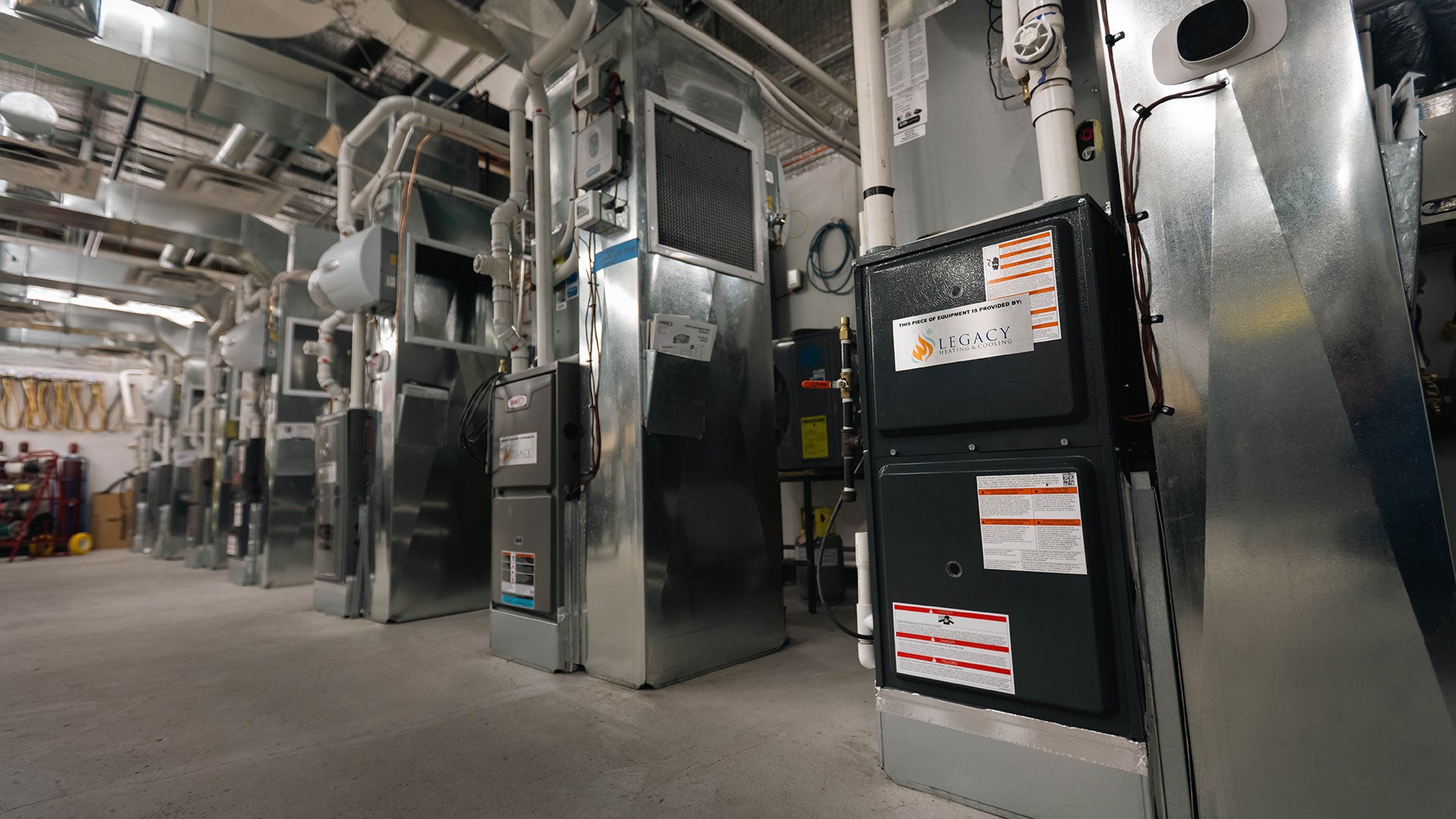 Row of large metal residential furnaces installed in a basement or utility room with exposed pipes and ductwork.