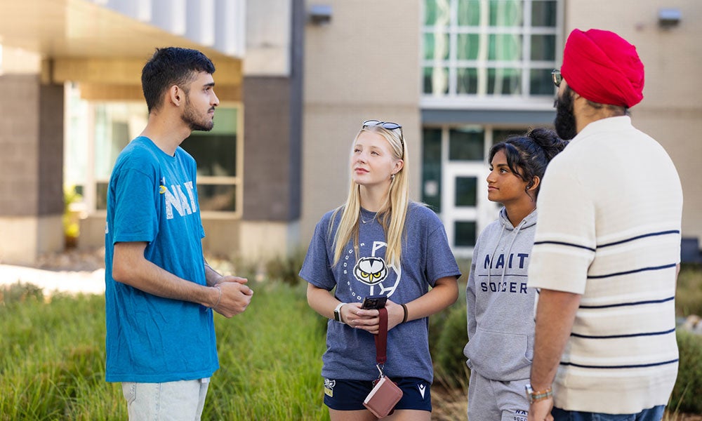 Four students standing outdoors on campus, talking together in front of an academic building.