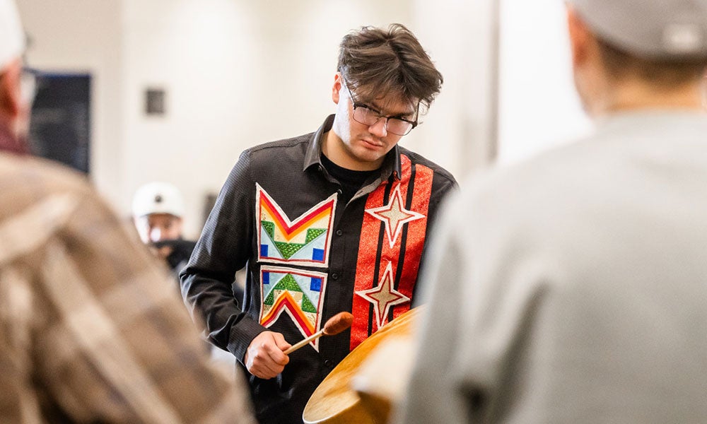 Person in a decorated shirt plays a hand drum indoors while other participants stand nearby in a gathering or cultural activity.