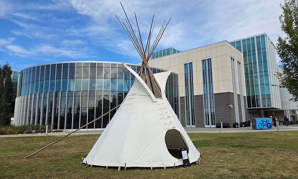 Completed white tipi standing on a grassy lawn outside a modern glass-and-concrete building under a blue sky.