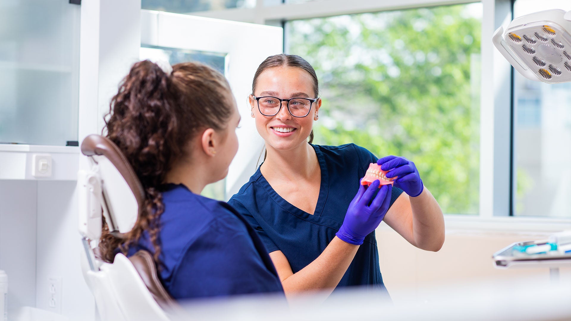 A female denturist technology student wearing blue scrubs and gloves showing a set of dentures to another student.
