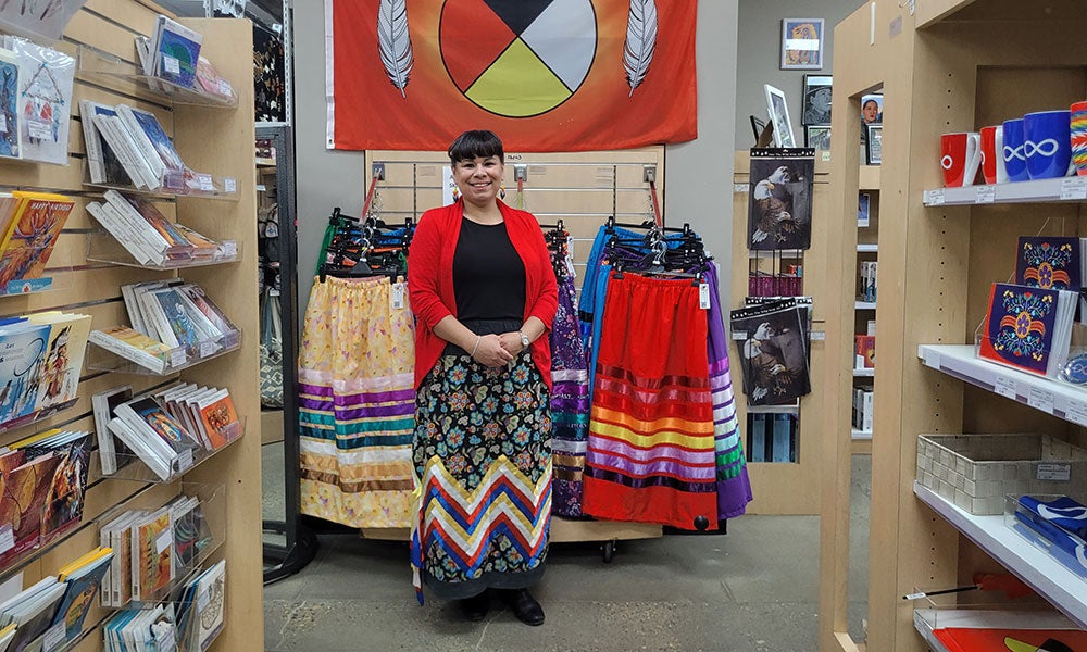 Person standing in a store between shelves of books and Indigenous-themed items, with colorful ribbon skirts displayed beneath an orange flag featuring the medicine wheel and feathers.