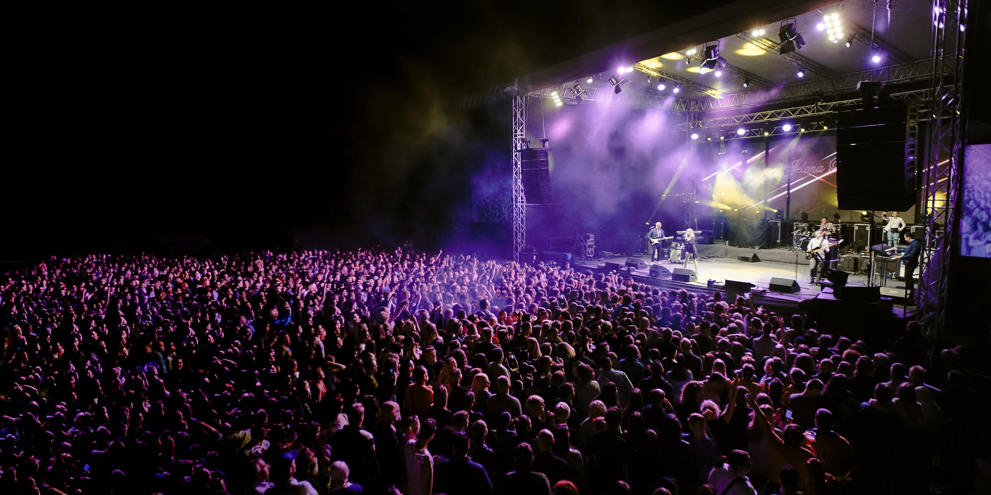 Large crowd gathered in front of an outdoor concert stage illuminated with purple and white lights during a live music performance at night.