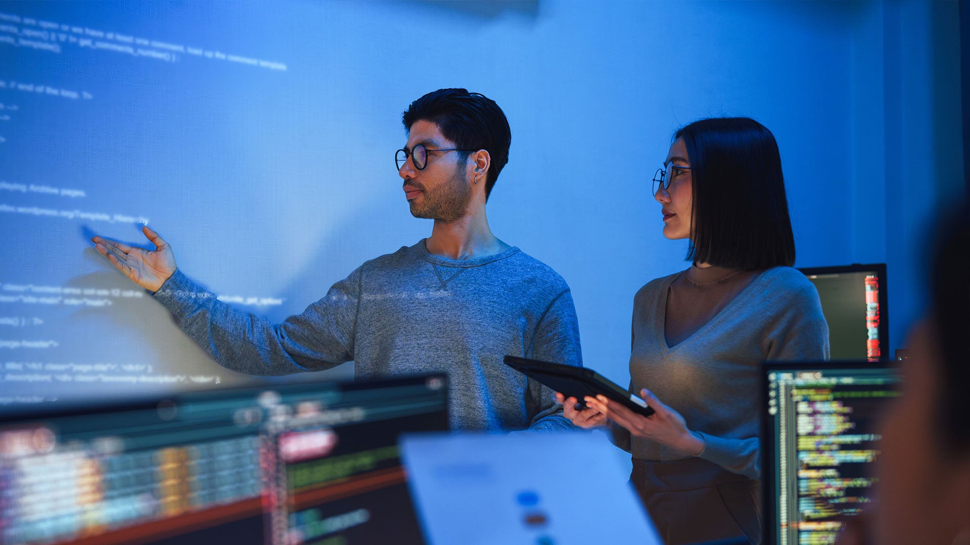 One person gesturing to a board displaying code, while another observes, holding a tablet. 