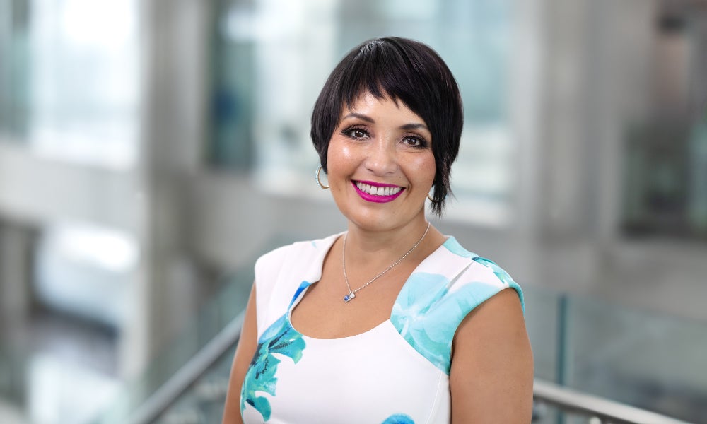 Head-and-shoulders portrait of a person with short dark hair smiling in a bright indoor office setting with a softly blurred background.