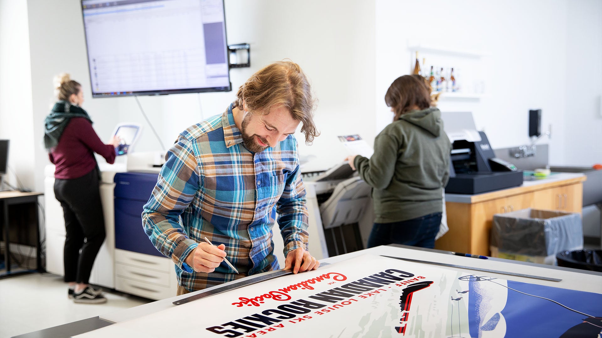 A designer working on a large, printed poster in a print production room, with two other people operating printers in the background.
