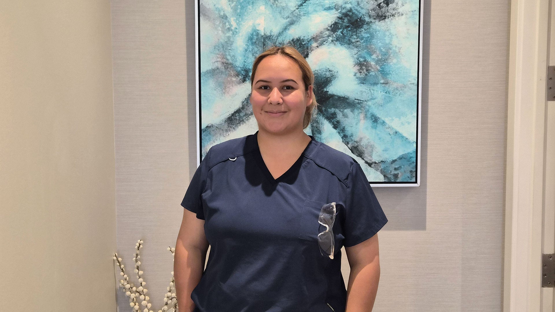 Person wearing dark blue scrubs standing indoors in front of an abstract blue painting on a beige wall.