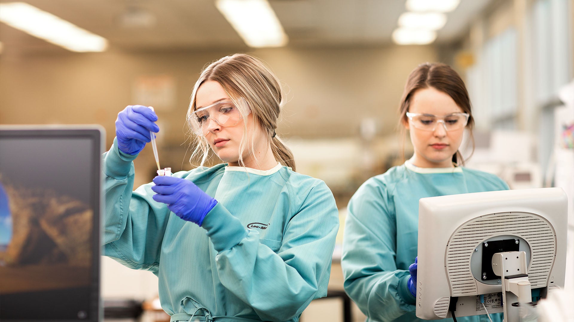 Two female students wearing gowns, safety glasses and gloves working in a lab.