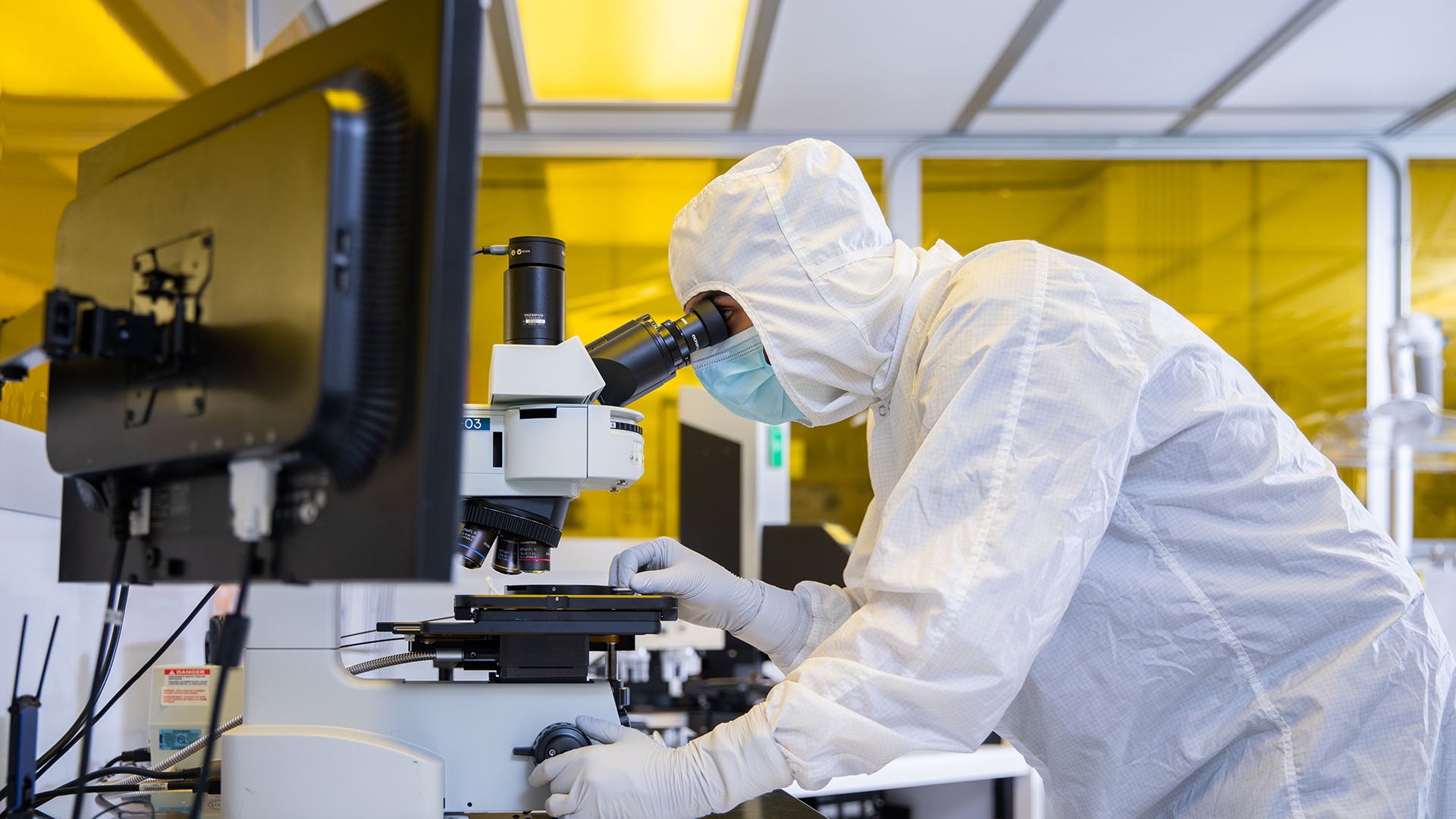 A learner in head-to-toe sanitation outfit looks at materials through a microscope in a lab setting.