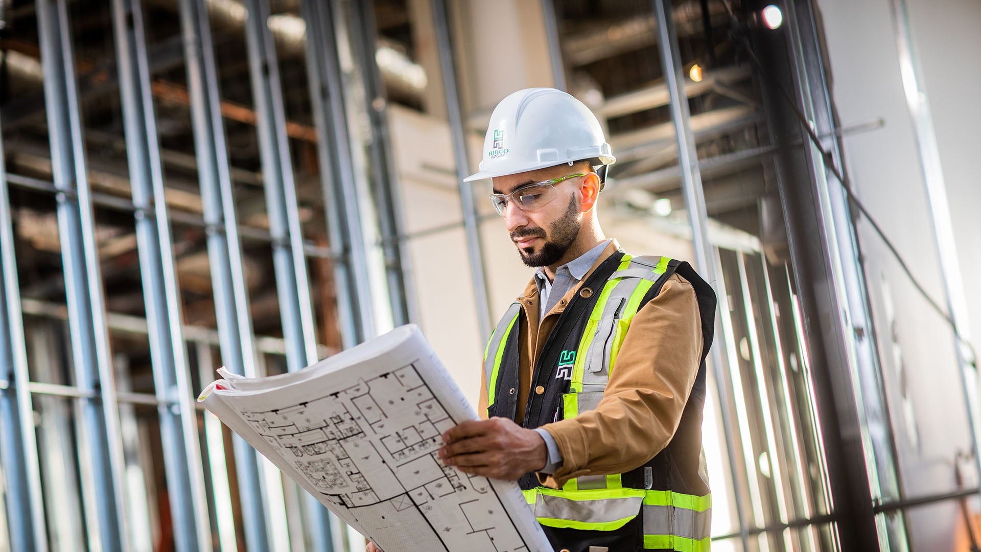 A project manager wearing a safety helmet, glasses, and vest, holding a blueprint on-site.