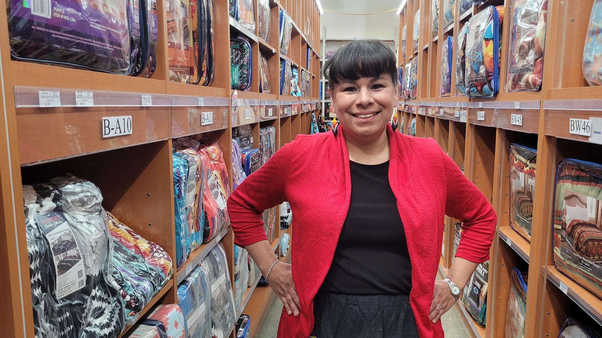 Person standing in a warehouse aisle lined with shelves of packaged blankets, wearing a red cardigan over a black outfit with hands on hips.