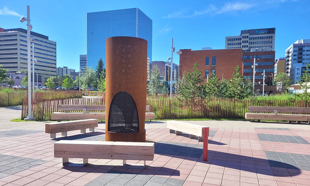 Urban plaza with benches surrounding a cylindrical public fire feature, framed by landscaped greenery and downtown office buildings under a bright blue sky.