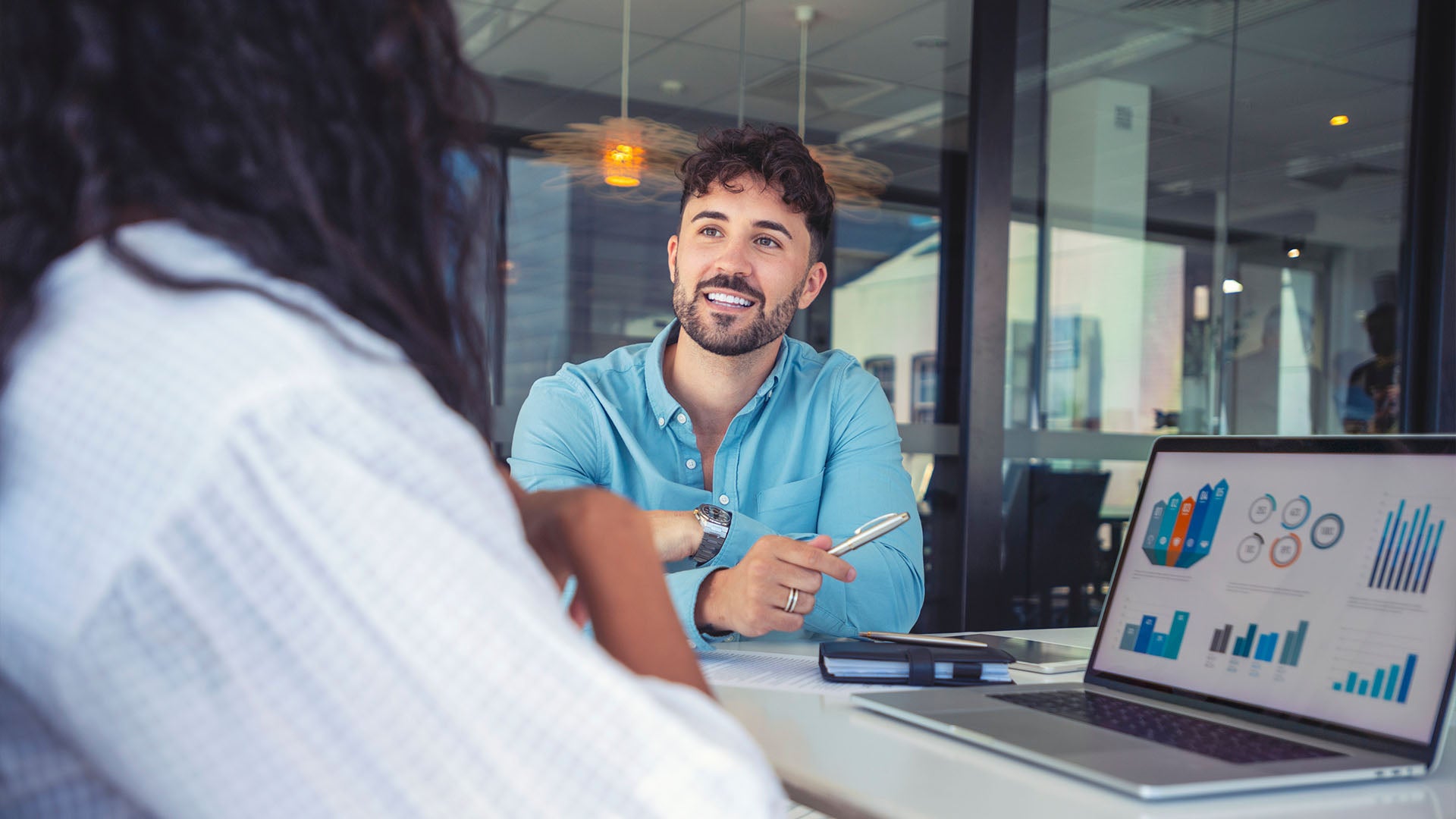 A man holding a pen smiles as he speaks with a woman seated in front of an open laptop.