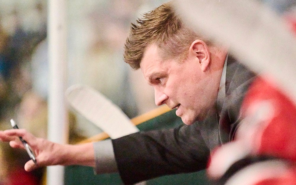 Individual seated behind a sports team bench, holding a pen and clipboard during an indoor hockey game, with players and arena activity visible in the foreground and background.