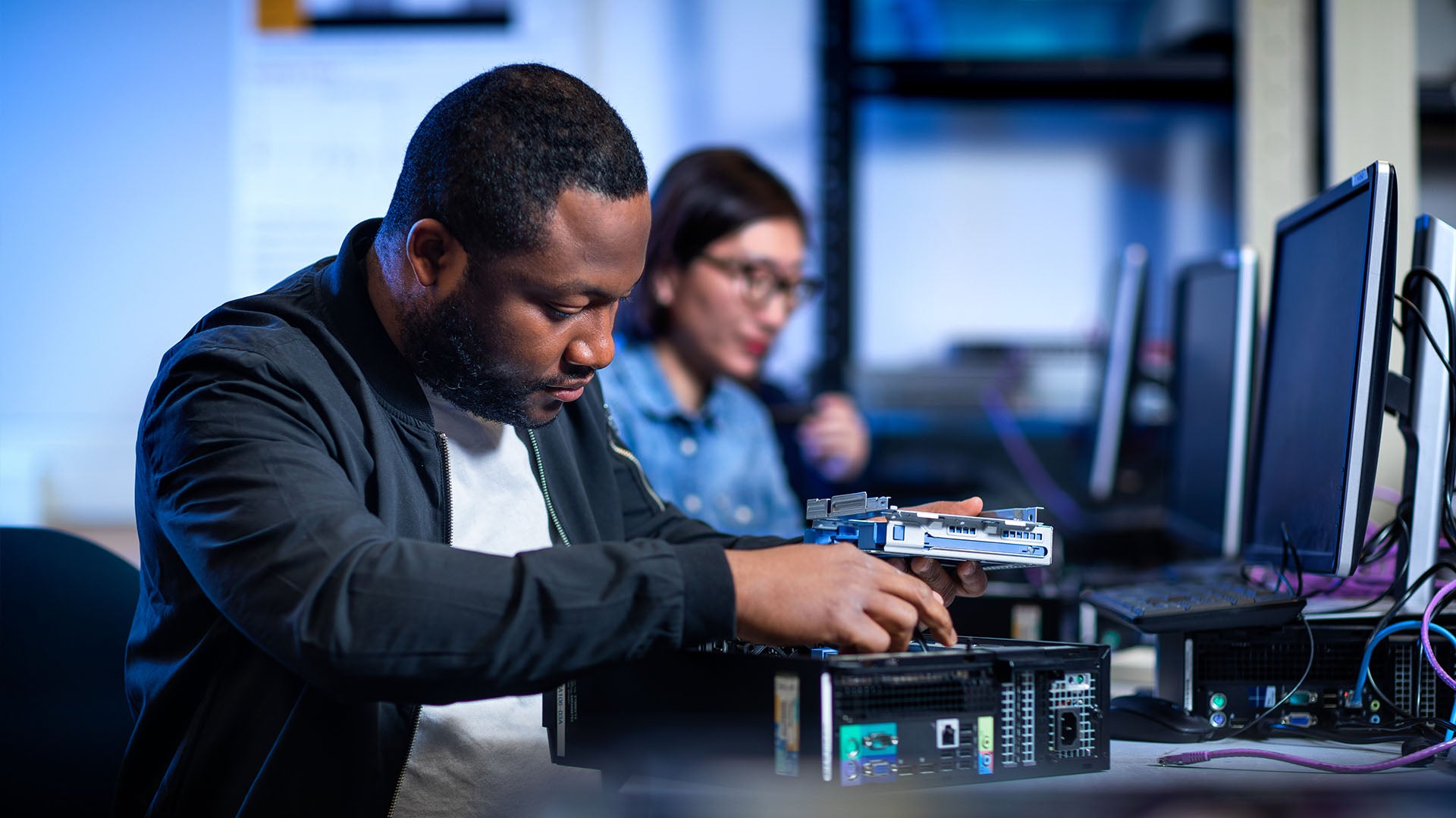 Man working on computer hardware in a lab, holding a component while another individual works on a computer in the background.