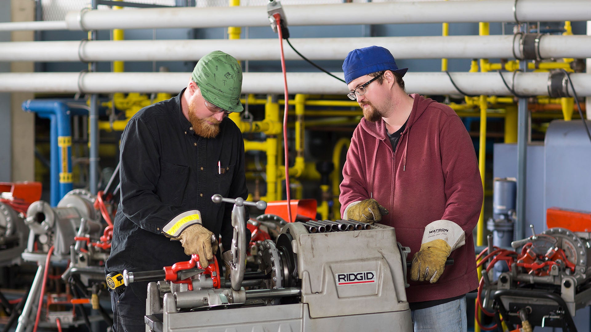 Two male steamfitter-pipefitter students wearing gloves operating a RIDGID pipe threading machine.
