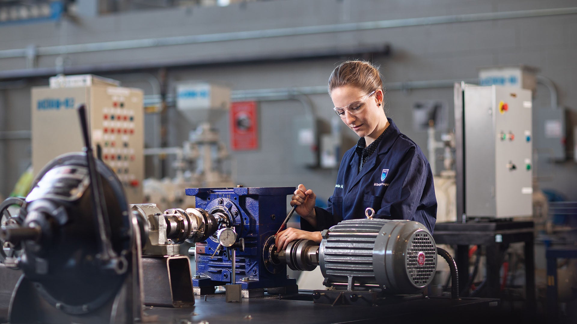 Female millwright wearing coveralls and safety glasses working in a shop.