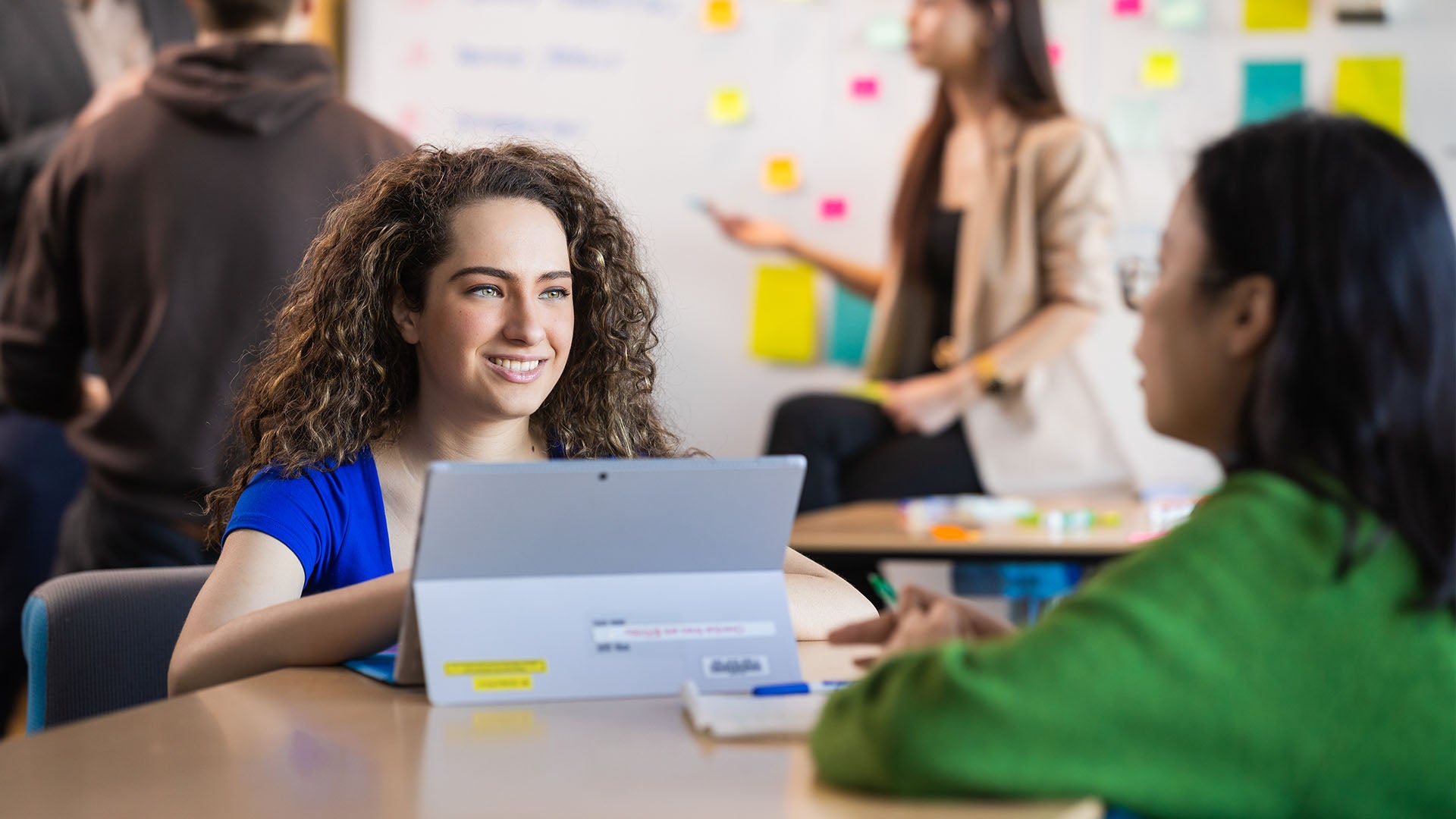 Two female students engaged in a discussion, with one working on a tablet. In the background, a group of students is gathered near a whiteboard covered with sticky notes, having a discussion.