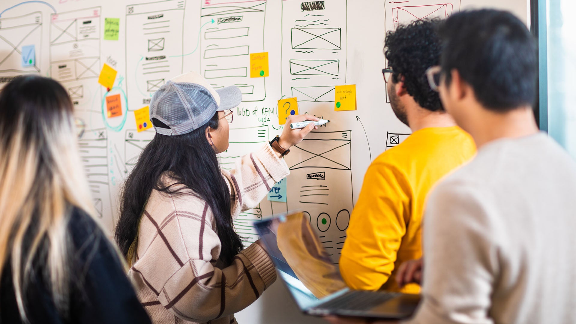 A group of four web design students standing in front of a whiteboard covered with wireframe sketches and colorful sticky notes. One student is actively writing on the board, while another holds a laptop.
