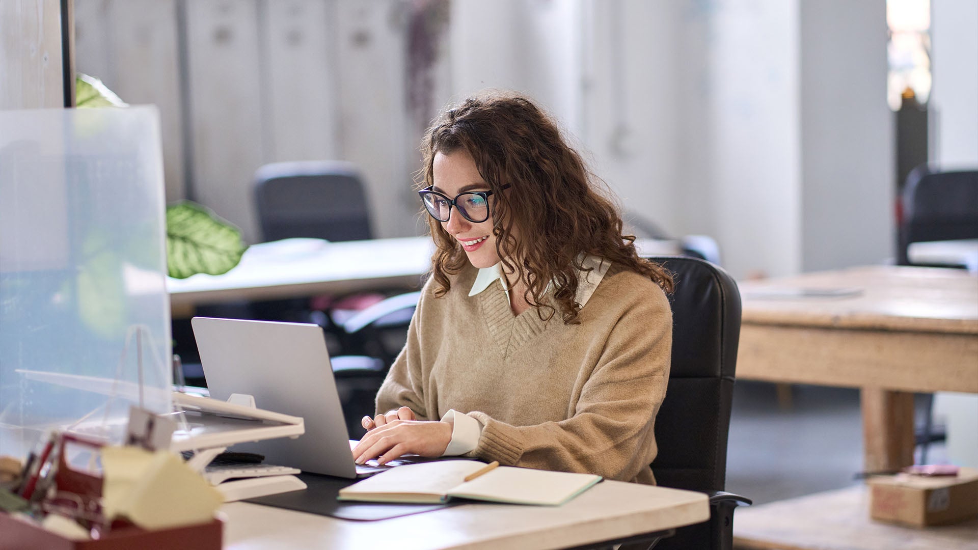 A cheerful woman wearing glasses and a sweater, working on a laptop at a desk with an open notebook in a bright office space.