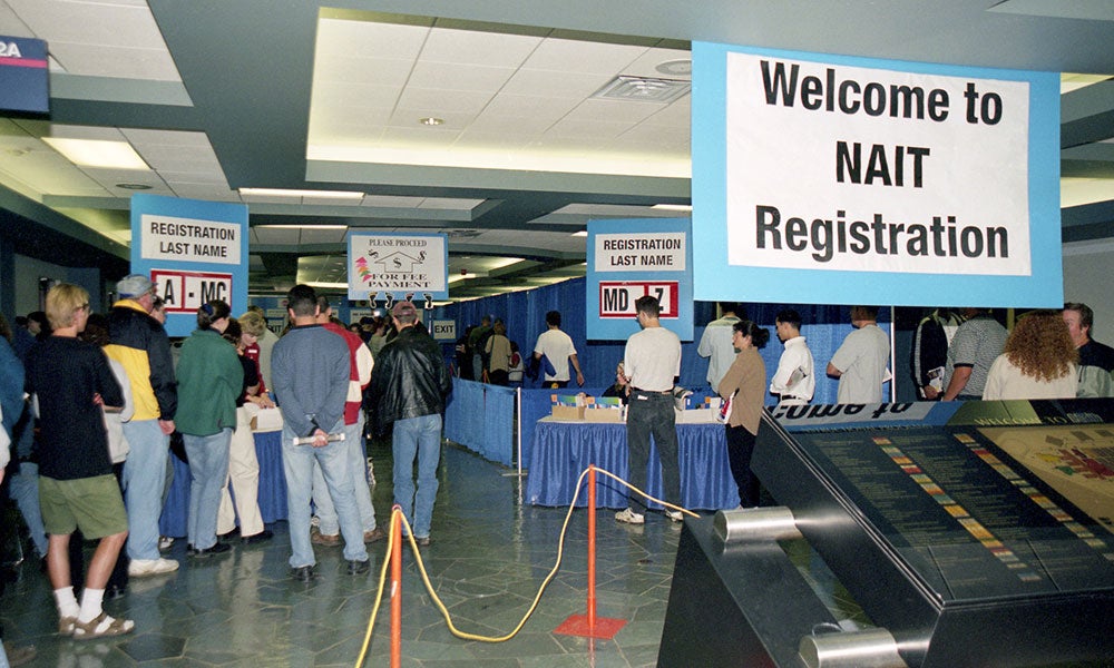 Registration event area with information tables, queue lines, directional signs, and visitors gathered beneath a large “Welcome to NAIT Registration” banner.