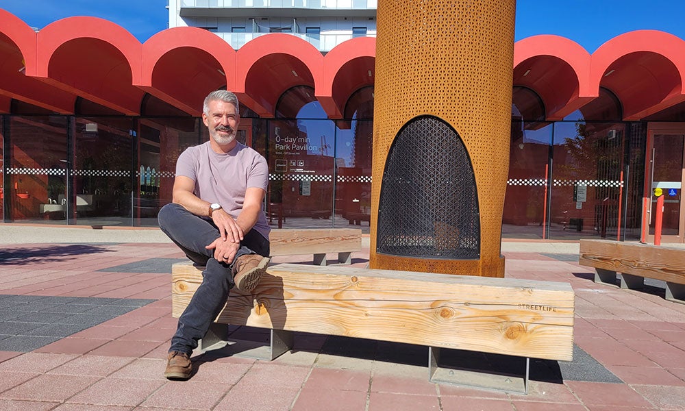 Person seated on a wooden bench in a sunlit public plaza, with a distinctive red-arched building facade and a tall cylindrical outdoor fireplace in the background.