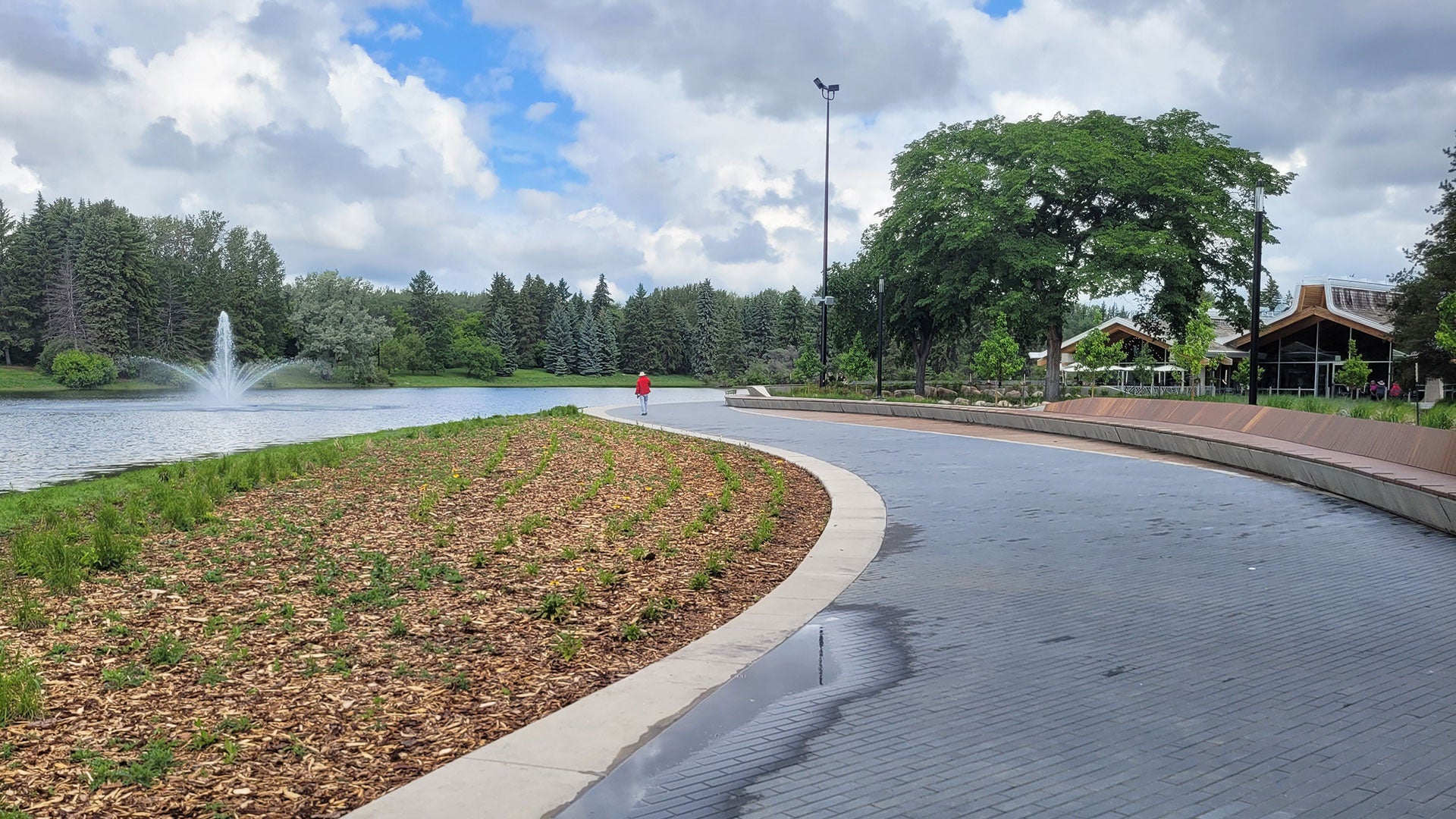 Curving waterfront path beside a lake and fountain, with trees, landscaped shoreline, and a cloudy sky in a park setting.