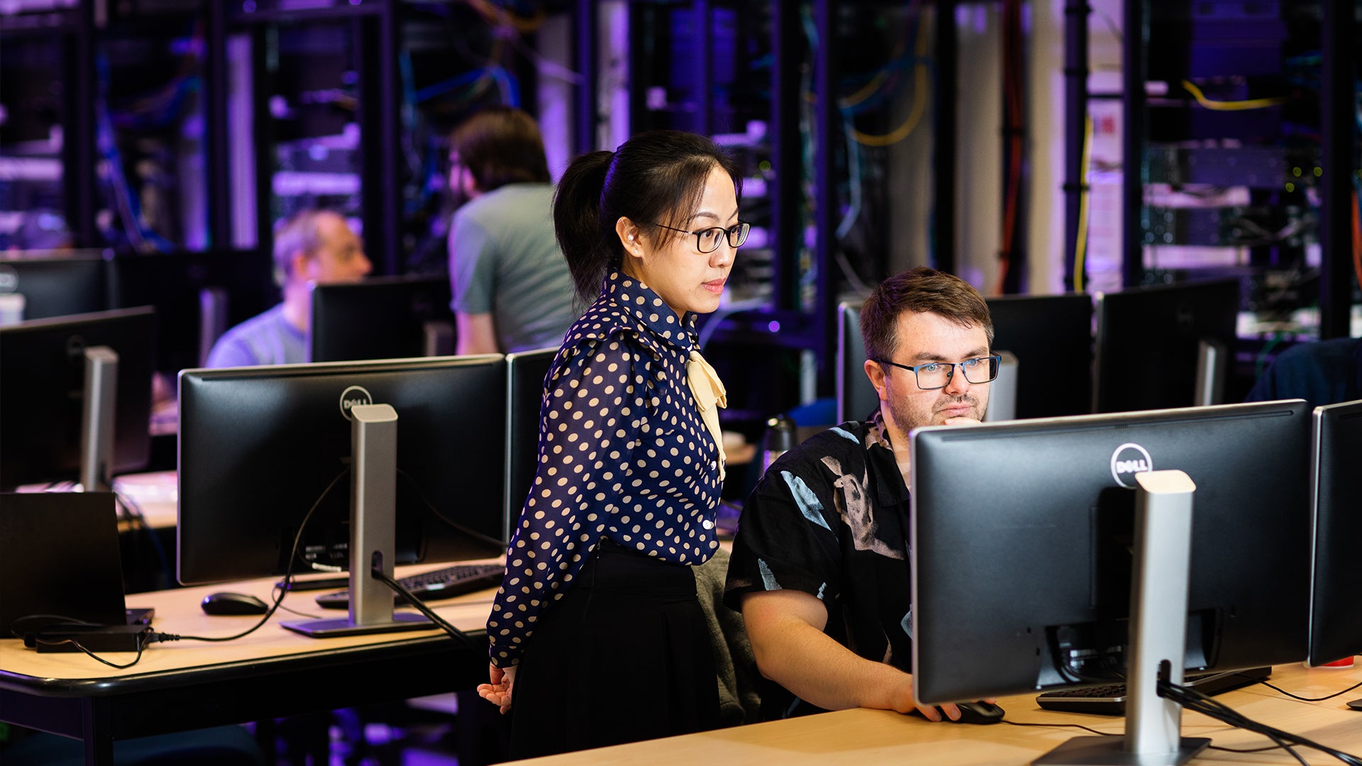 A male network engineering technology student works on a desktop computer in a classroom, while a female instructor stands and observes. The room contains other computers and students working.
