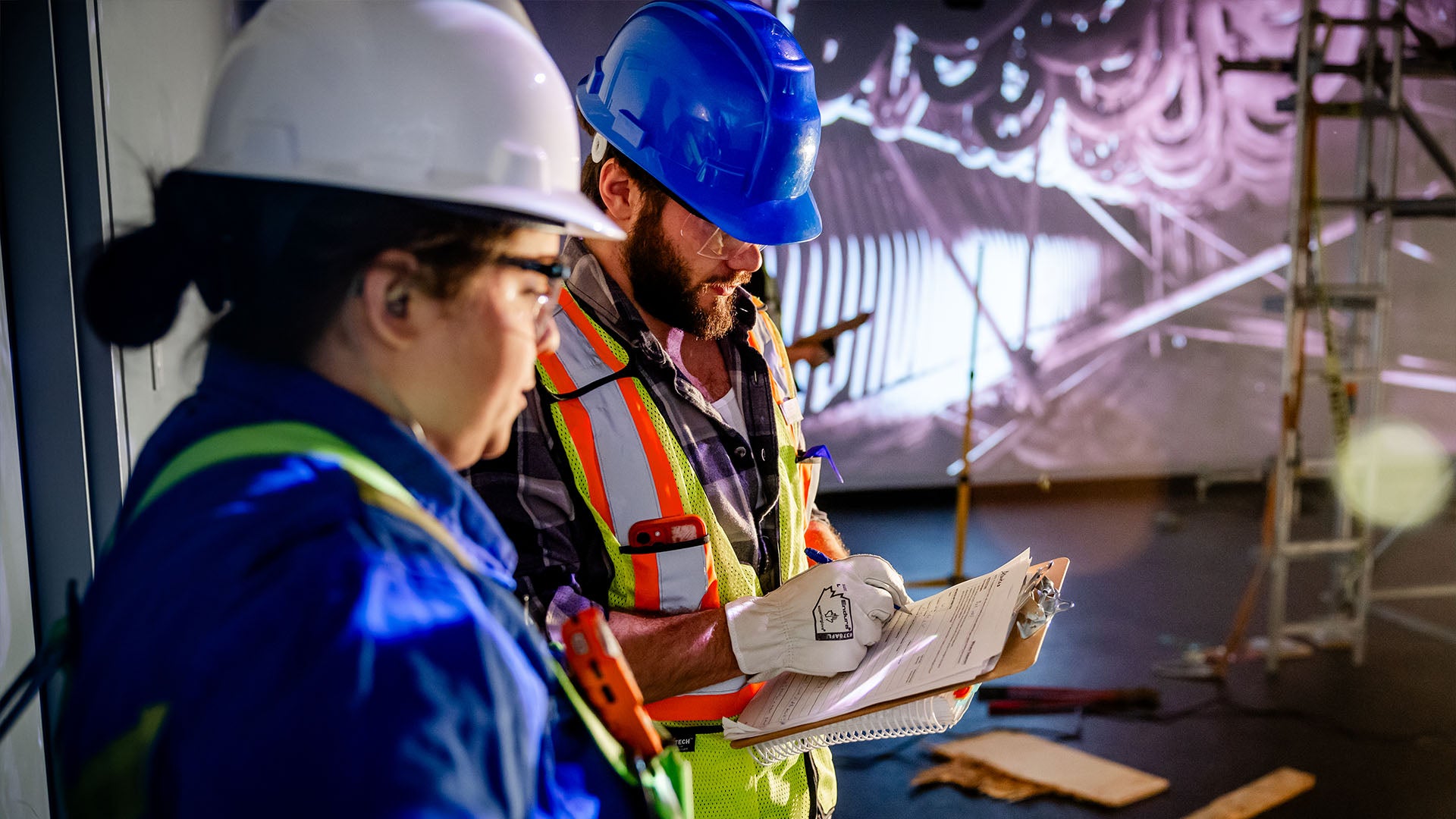 Two students in hard hats during a health and safety simulation: a female student in coveralls and a male student in a high-visibility vest and gloves, writing on a clipboard.