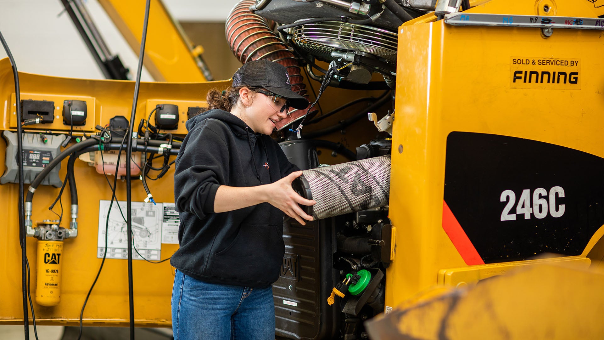 A woman wearing safety glasses and a cap working on the engine of a skid steer loader in a workshop setting.