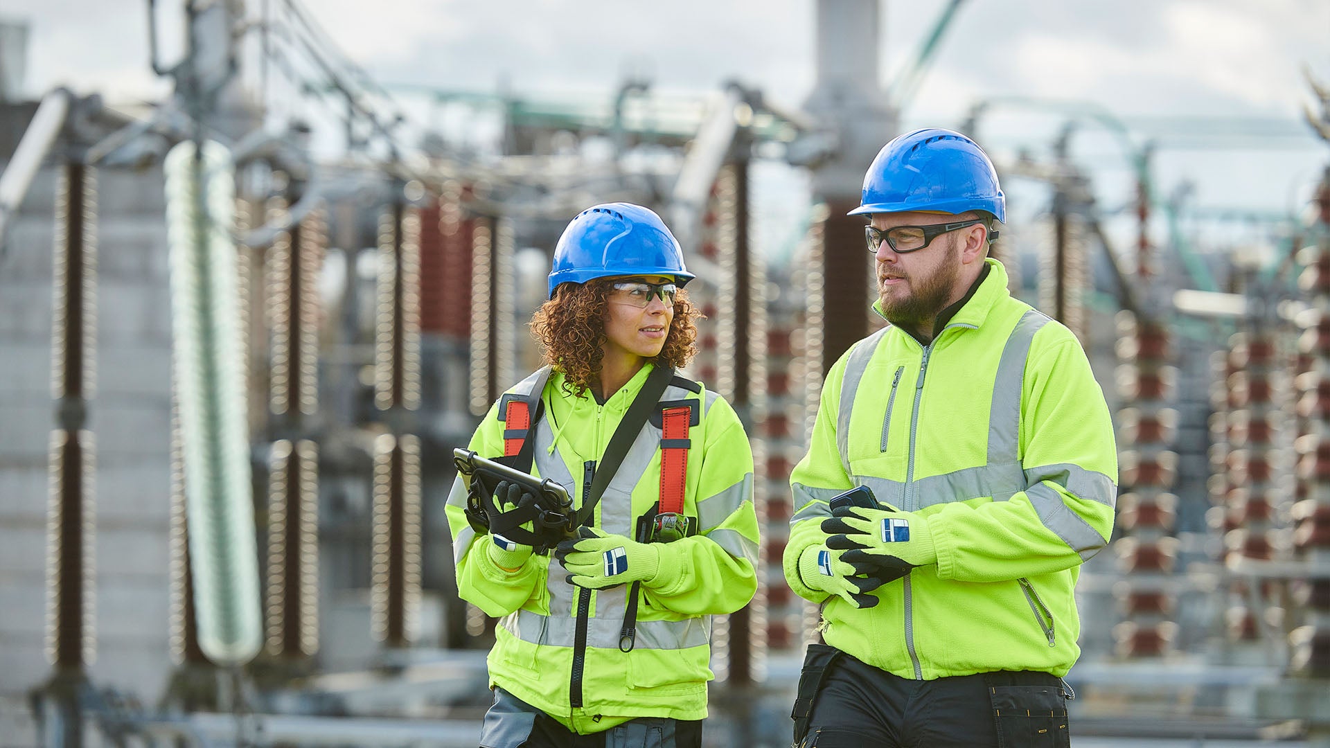 Two individuals wearing high visibility jackets, safety glasses and hard hats standing outside.