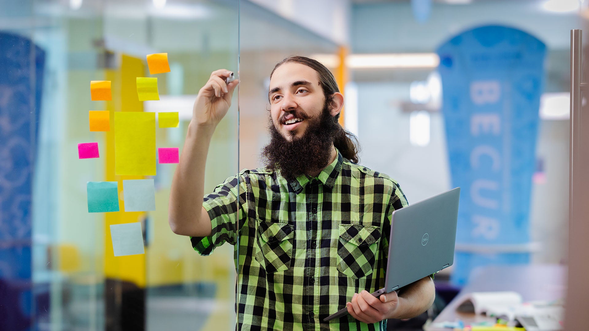 A man uses a marker to draw on a glass wall covered with sticky notes while holding a laptop during a planning session.