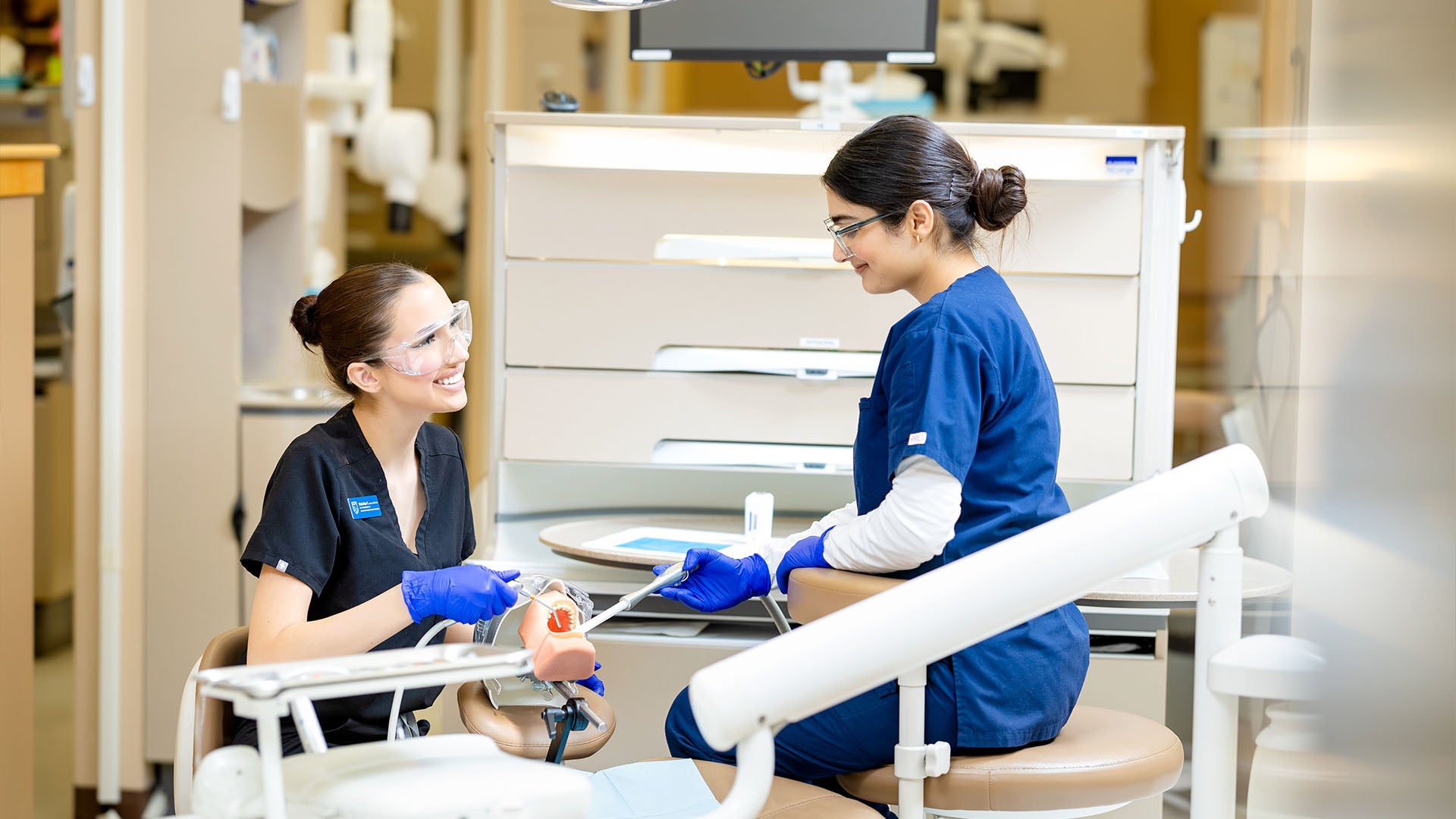 Two women wearing blue scrubs practice using dental cleaning tools on