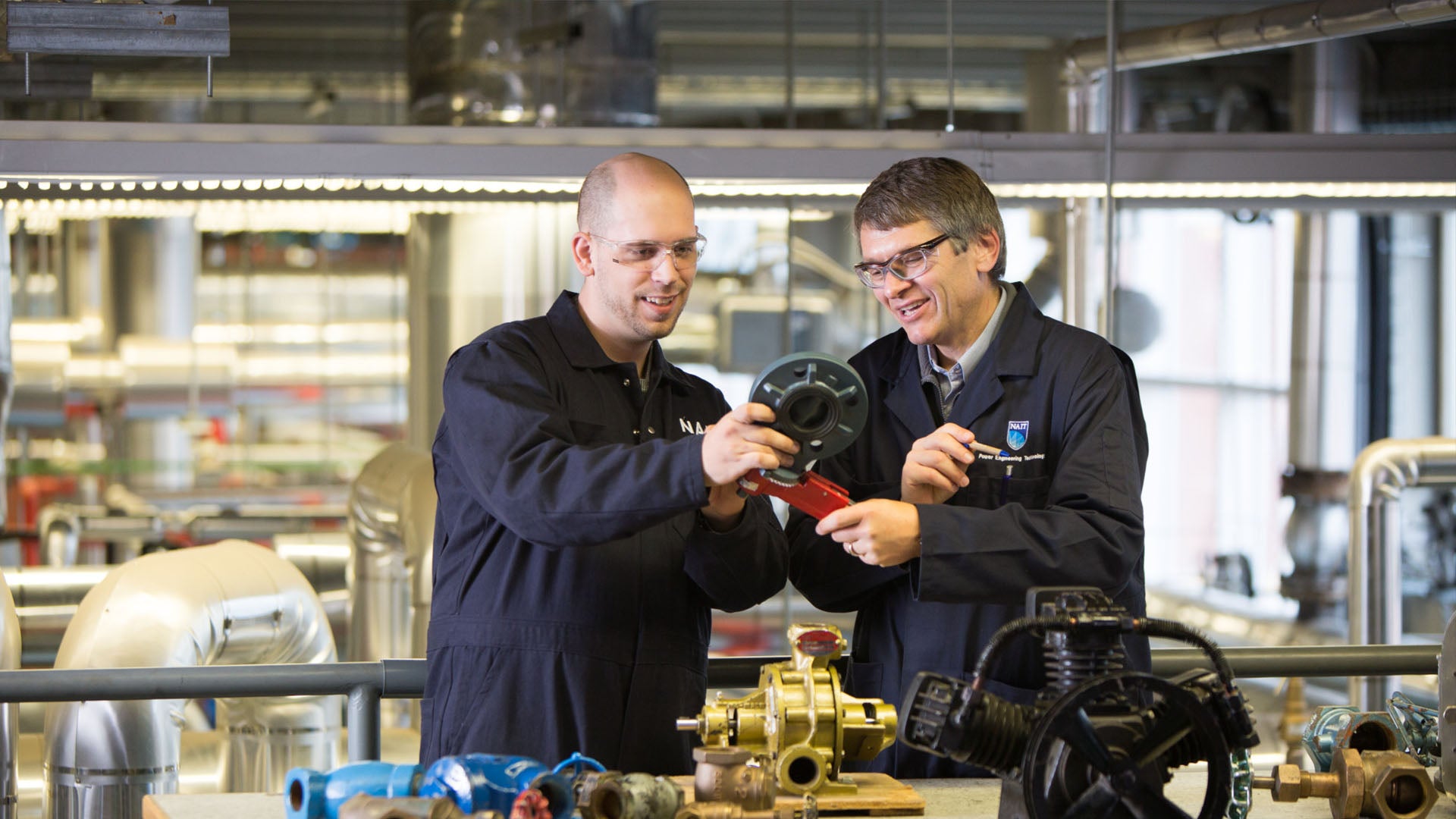 Two power engineers wearing coveralls and safety glasses in a workshop.